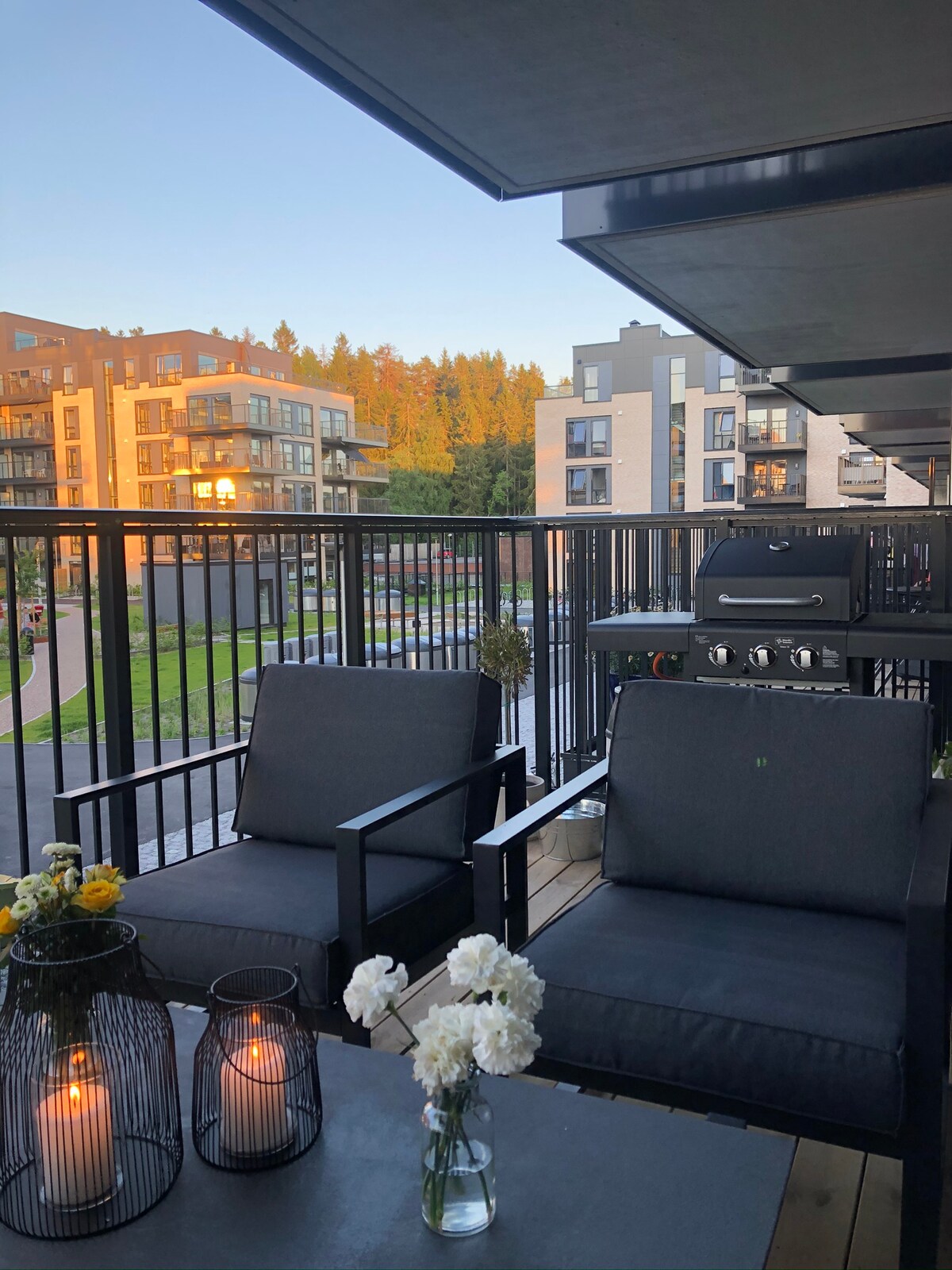 A balcony area features two dark, cushioned chairs and a small table adorned with candles and flowers. A grill is positioned on one side. In the background, modern apartment buildings and trees are visible, illuminated by warm evening light.