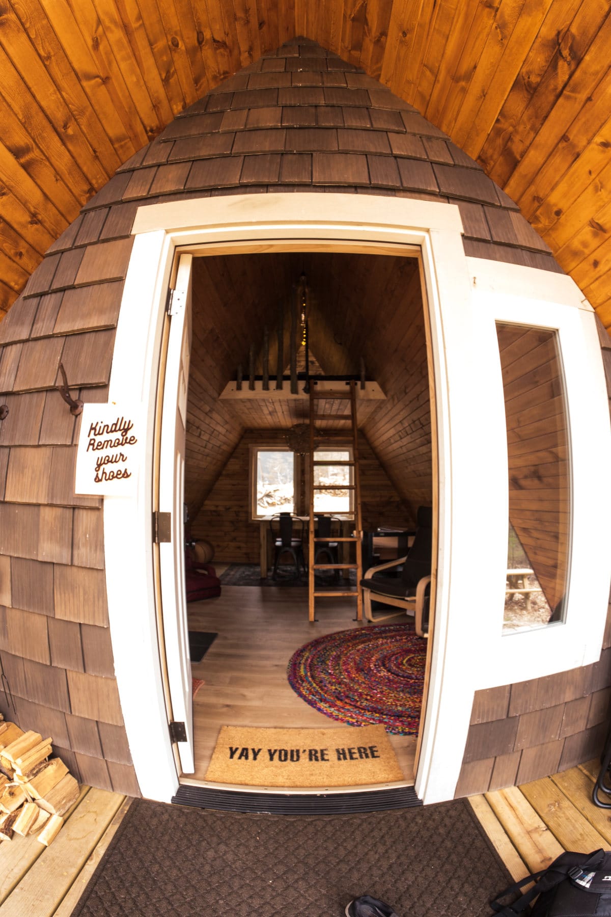The entrance of the A-Frame cabin features wooden shingle walls and a cozy welcome mat. A circular, colorful rug sits on the floor, and a ladder leads to the loft. Natural light streams through large windows, inviting guests into the rustic interior.