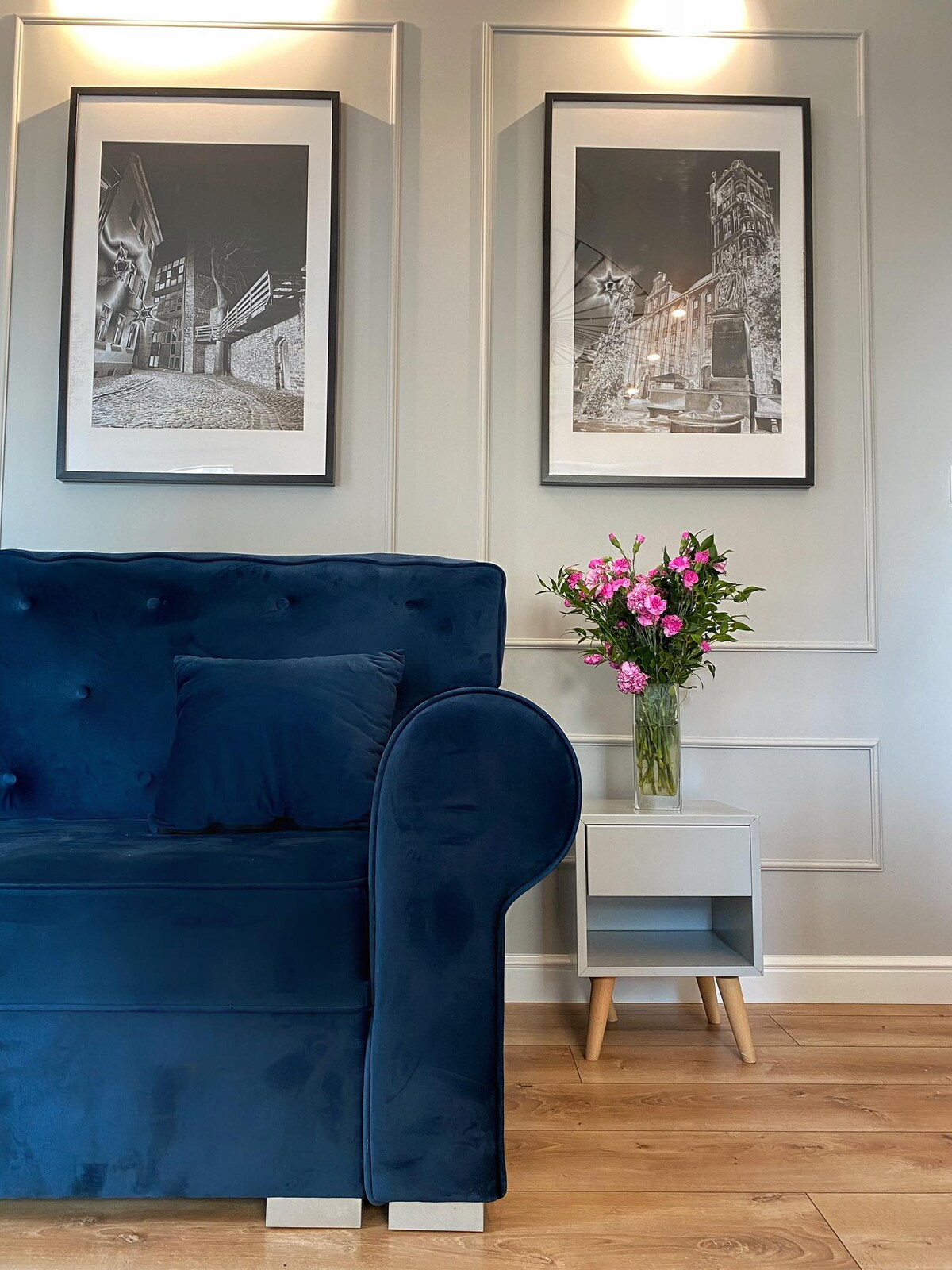 A navy blue sofa is positioned against a light grey wall adorned with two framed black-and-white photographs. A small table beside the sofa holds a vase of pink flowers, adding a touch of color to the serene setting.