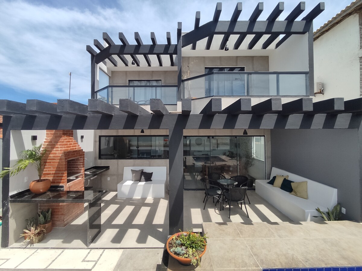 The exterior features a contemporary two-story house with large glass doors, enhancing the connection to the outdoor space. A covered patio includes stylish seating and a dining area. A brick barbecue is positioned nearby, surrounded by greenery and potted plants, providing a relaxing atmosphere.