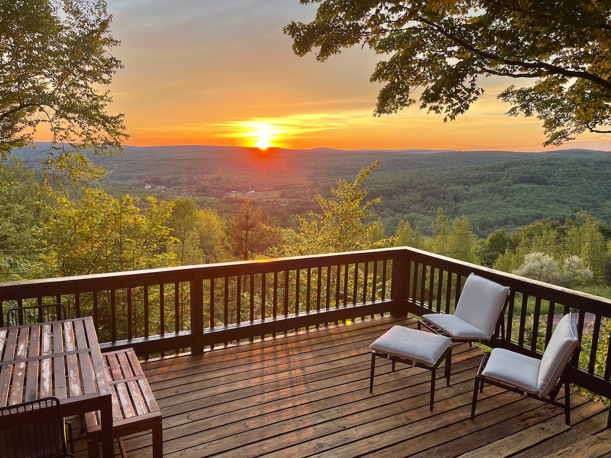 A spacious wooden deck overlooks a scenic landscape, framed by lush trees. Two lounge chairs are positioned beside a wooden table, with a stunning sunset casting warm hues across the valley below. The expansive view captures the tranquility of the surrounding nature.
