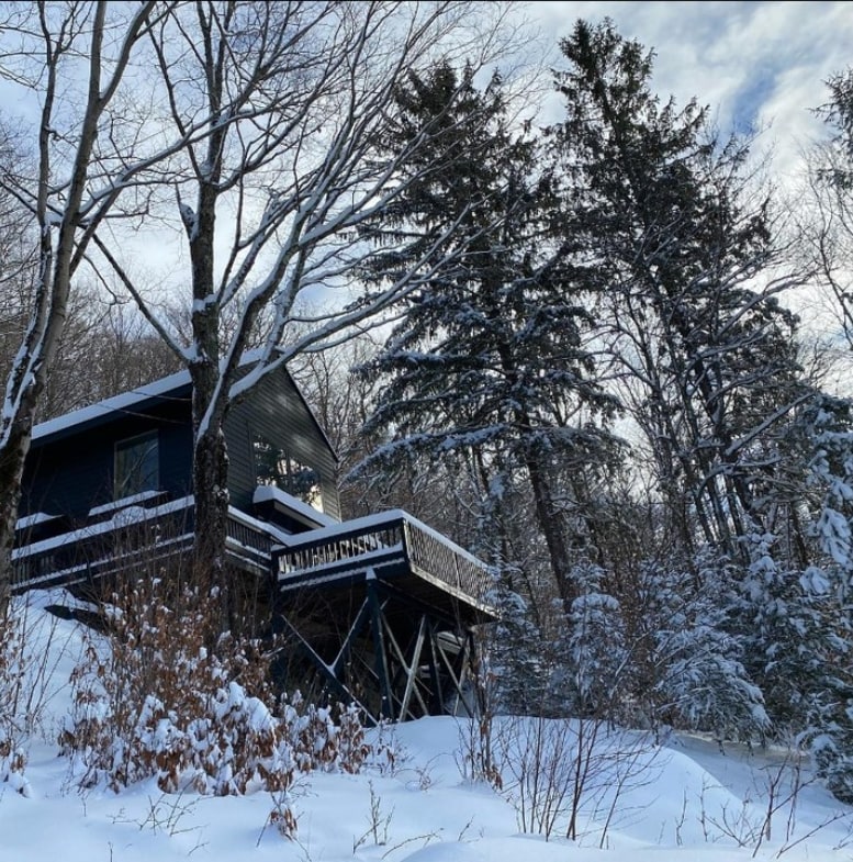A modern house nestles among snow-draped trees on a hillside. The structure features a prominent deck supported by stilts, framed by the serene winter landscape. Snow blankets the ground, and the scene is enhanced by the blue tones of the house contrasting with the white snow.