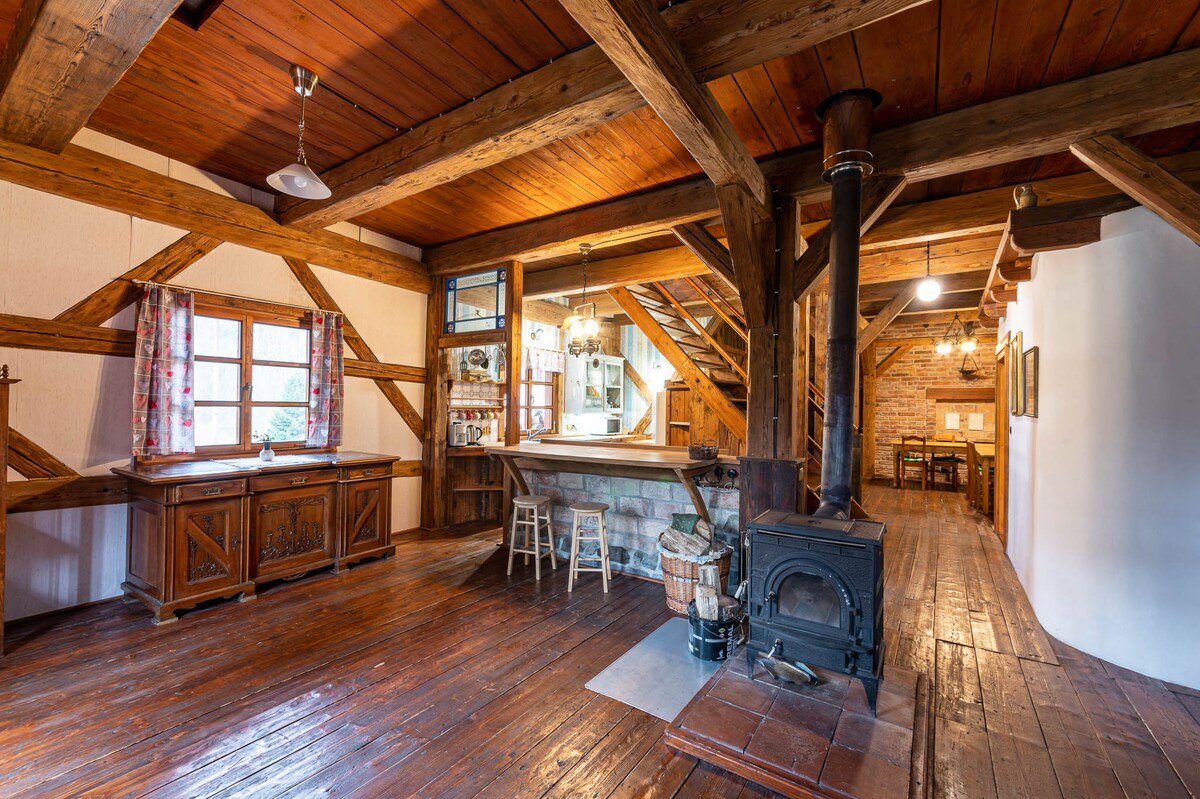 An open living area features exposed wooden beams and rustic wood flooring. A wood-burning stove is positioned next to a bar area with stools. Natural light streams through windows adorned with patterned curtains, illuminating the inviting space.