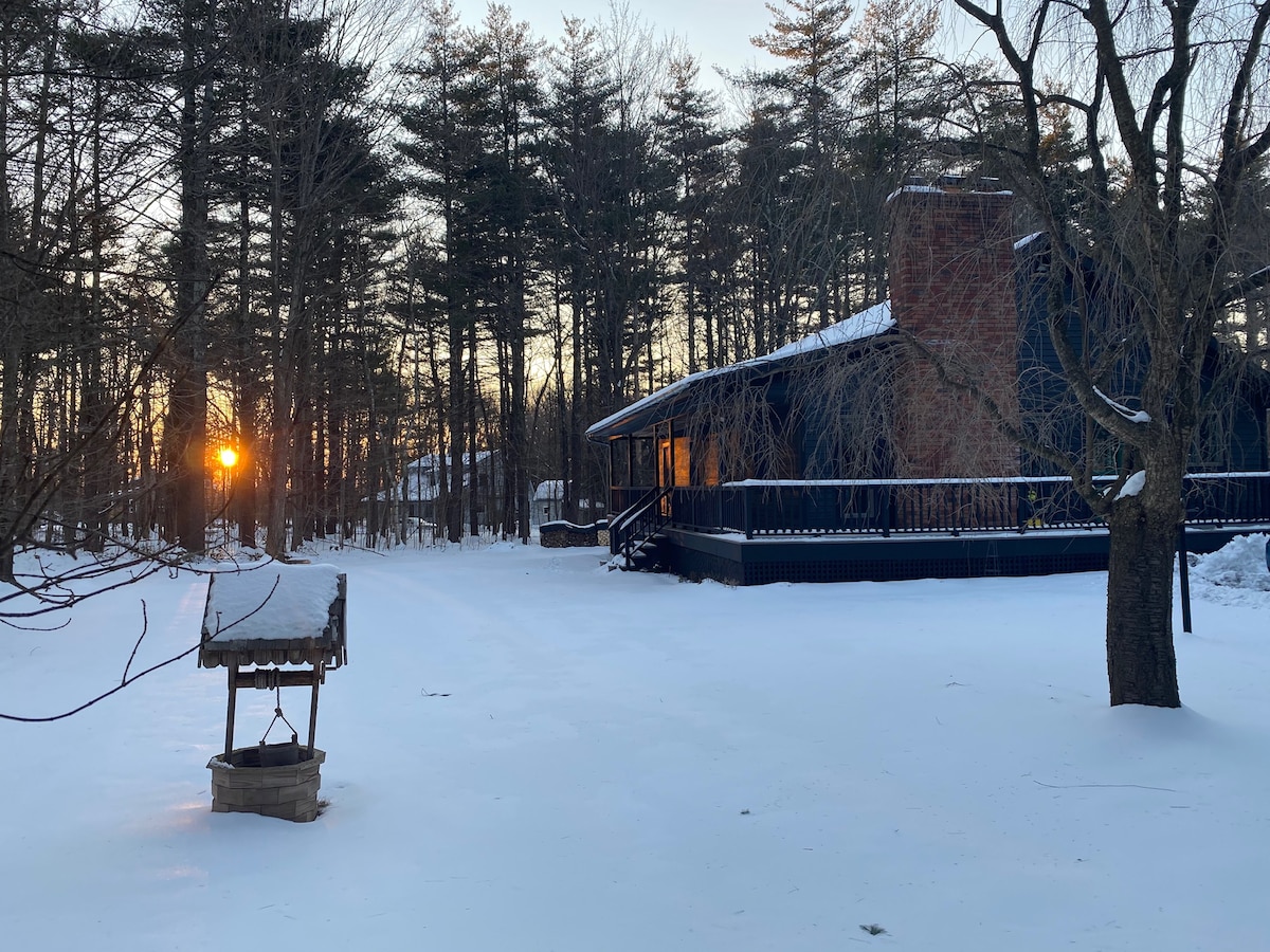 The exterior of the home is set against a winter landscape, with a layer of fresh snow covering the ground. A rustic well is positioned in the foreground, while the sun is seen setting behind the trees, casting a warm glow on the house.
