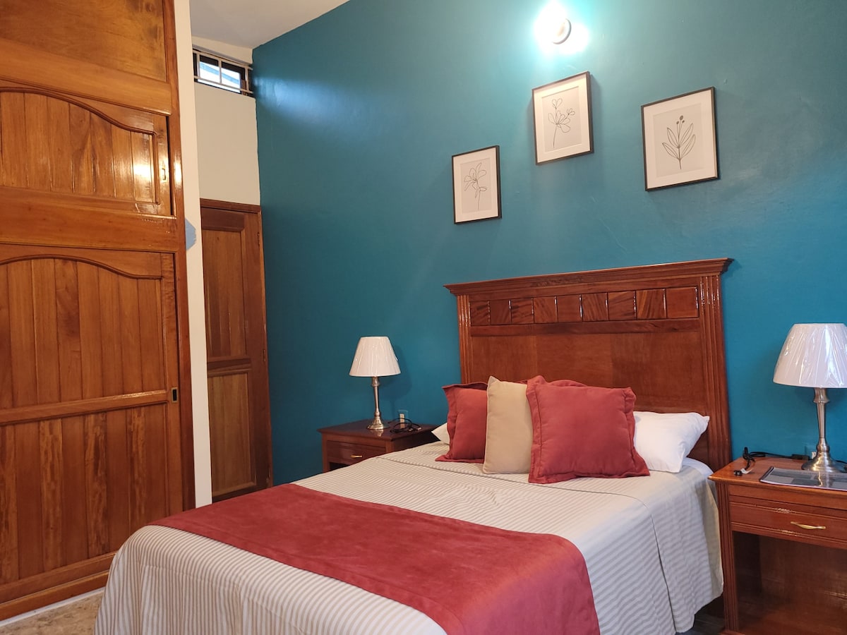 A modern bedroom features a spacious bed adorned with neutral bedding and accent pillows. Two bedside tables hold elegant lamps, while framed botanical prints decorate the vibrant blue wall. A wooden closet and warm timber accents enhance the room's inviting atmosphere.
