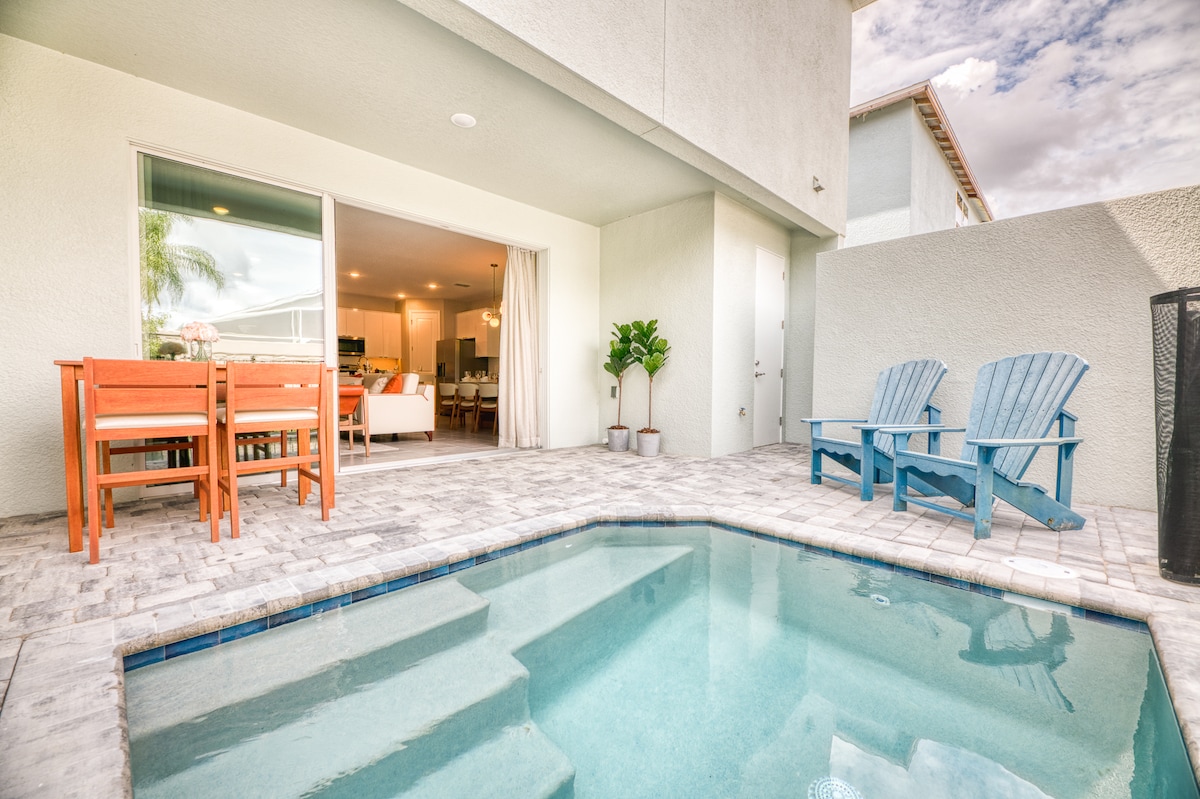 A private pool is featured in the backyard, surrounded by a stone patio. Outdoor seating is provided with a dining table and chairs, alongside two blue lounge chairs. Large glass doors lead into the interior space, allowing natural light to flow indoors.
