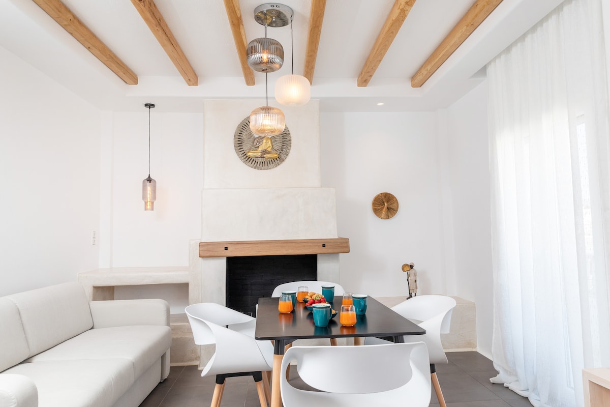 A light-filled living area features a modern sofa alongside a dining table set with colorful dishes. Pendant lights hang above, and a decorative fireplace adds warmth to the space. Sheer curtains allow soft natural light to enter, complementing the wooden ceiling beams.
