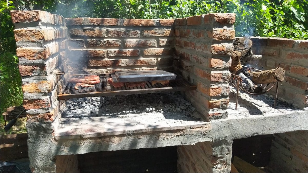 A traditional outdoor grill is captured with bricks forming a sturdy structure. Smoke rises from the grill, where food is being prepared over glowing coals. Nearby, a section for firewood is visible, alongside a natural green backdrop.