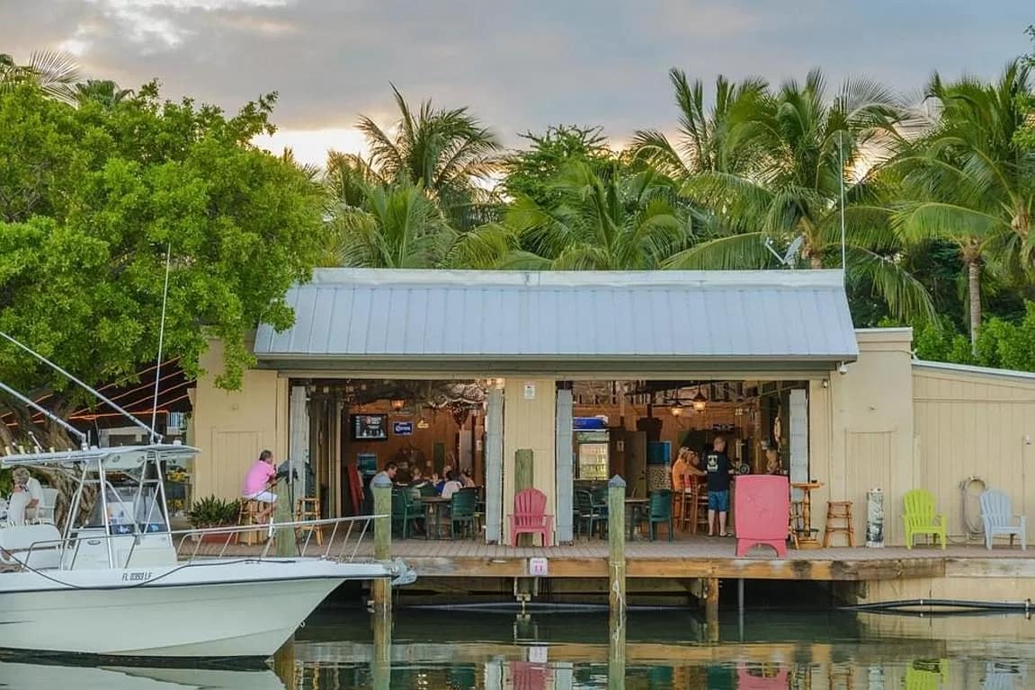 A waterfront bar and grill setting features a wooden deck lined with colorful adirondack chairs. Palm trees provide greenery and shade. The entrance reveals diners inside, enjoying the relaxed atmosphere, with boats docked nearby reflecting in the calm water.