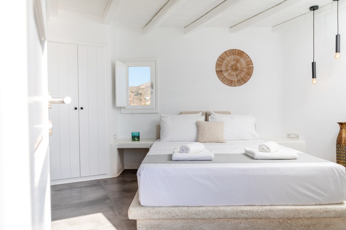 A serene bedroom features a spacious bed adorned with fresh linens and neatly arranged towels. Natural light filters through an open window, illuminating the light-colored walls and simple decor. A wooden accent piece and a minimalist closet complement the calming atmosphere.