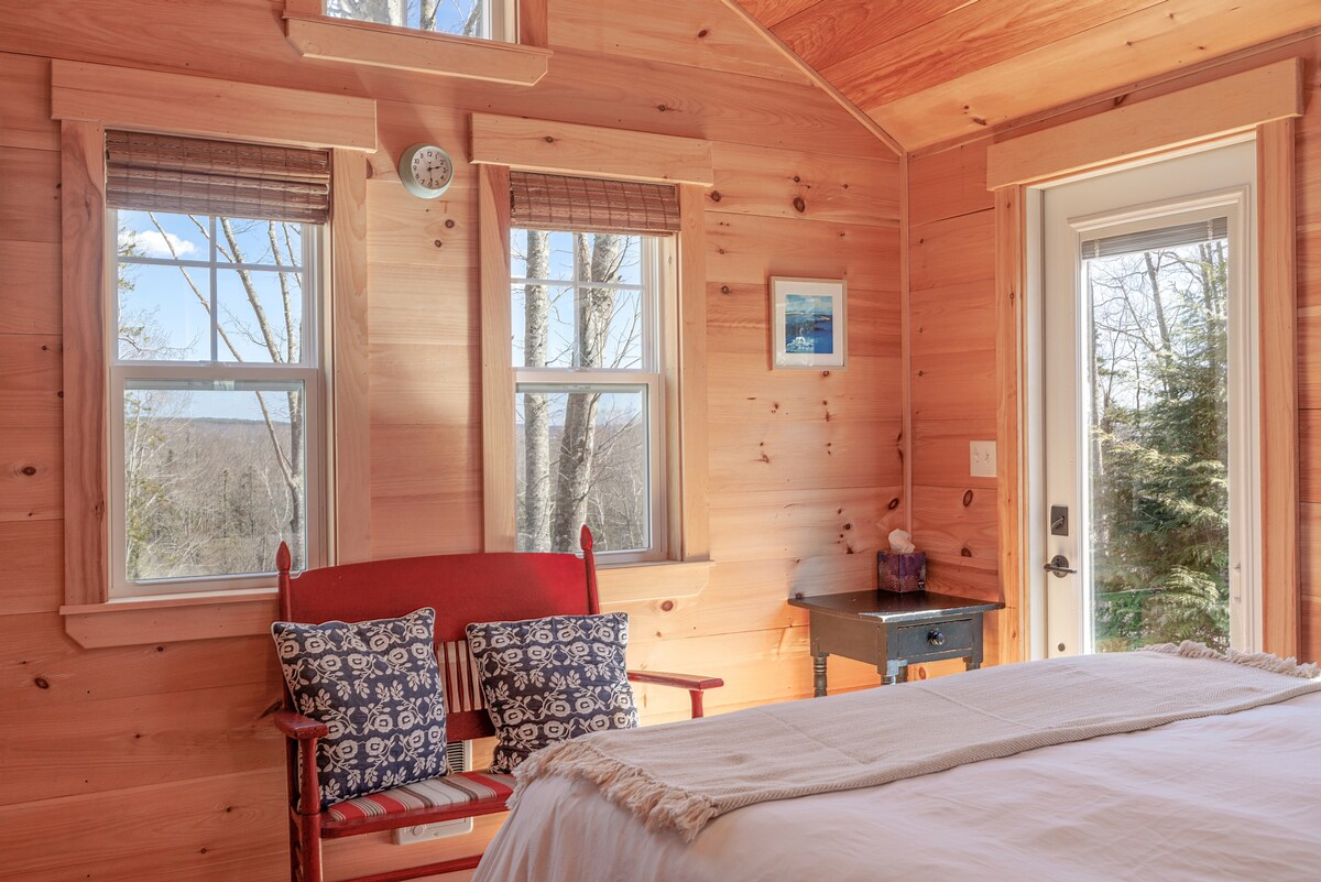 The interior of the cozy cabin features wood-paneled walls and a queen bed draped with a soft blanket. A small table with a lamp sits beside a red bench adorned with patterned cushions. Natural light fills the space through multiple windows, offering views of the surrounding forest.