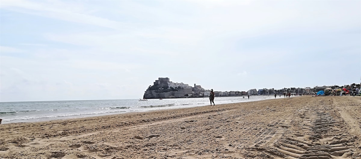 A sandy beach is depicted, with gentle waves lapping at the shoreline. In the distance, a historic castle, sitting on a rocky outcrop, overlooks the water. Footprints are visible in the wet sand, and some beachgoers can be seen strolling along the shore.