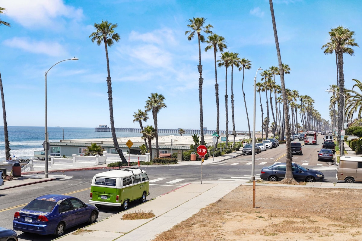 A scenic view captures palm trees lining the street, leading towards the ocean and a pier in the distance. A variety of parked vehicles, including a green van, are visible along the road. The sky is bright and clear, enhancing the coastal atmosphere.