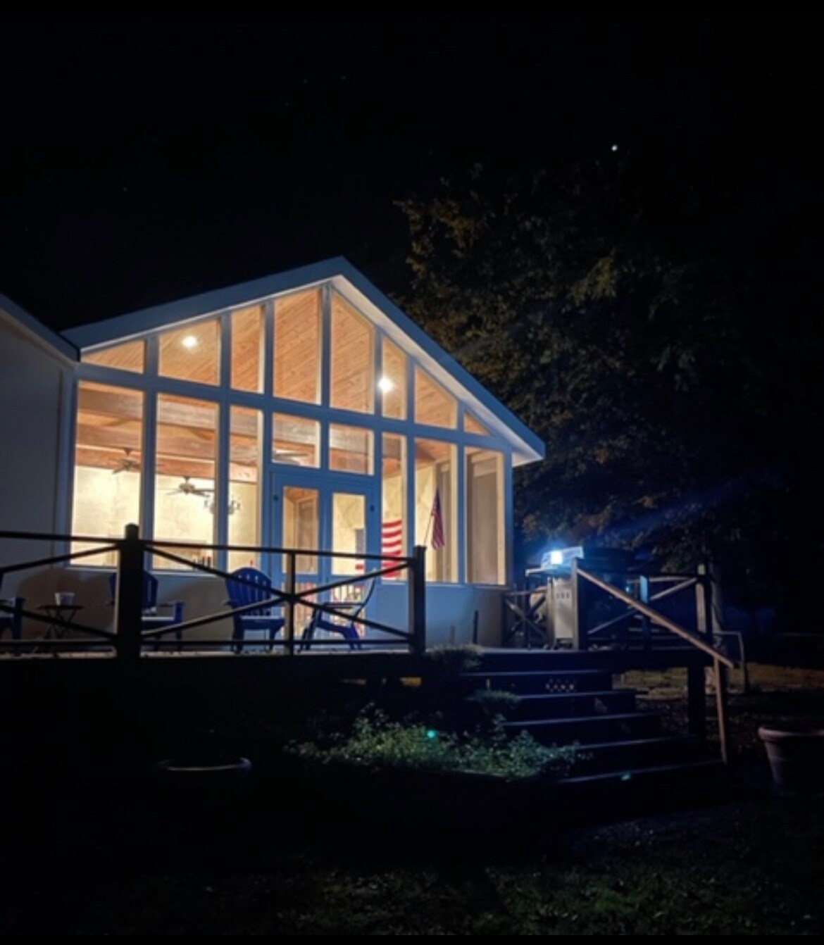 The exterior of the house is featured at night, showcasing large glass windows illuminated by warm indoor lights. A welcoming porch with seating is visible, and a staircase leads to the entrance. The surrounding area is dark, creating a contrast with the bright facade.