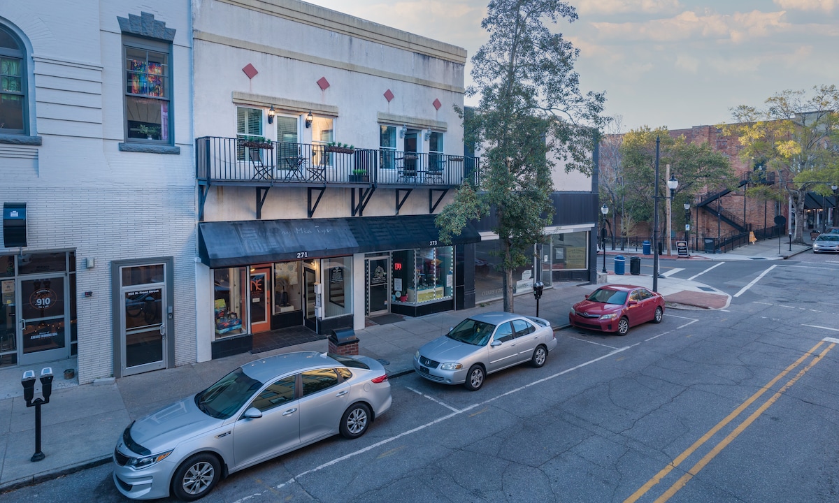 The exterior view presents a historic building with a balcony overlooking Front Street. Below, storefronts line the street, and parked cars are visible along the curb. The surrounding trees and sidewalk create a welcoming city atmosphere.