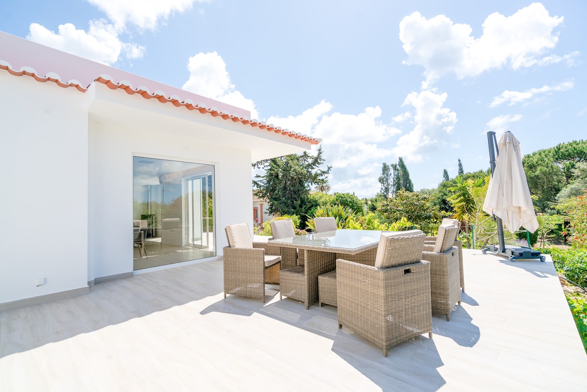 An outdoor terrace features a large dining table surrounded by six woven chairs under a sunshade. The expansive space is designed for al fresco dining, with greenery visible in the background enhancing the natural setting.