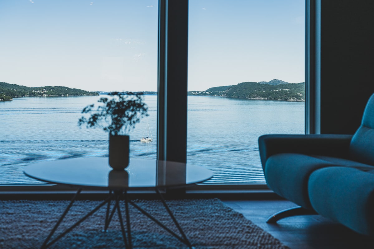 A spacious living area is framed by large windows offering stunning views of the serene water and surrounding hills. A coffee table with a small vase of flowers is situated in the foreground, enhancing the peaceful setting.