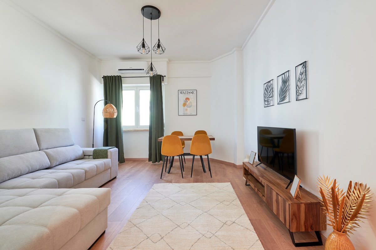 The living area features a light grey sectional sofa and a wooden TV stand. A round dining table with two orange chairs sits nearby. Large windows with green curtains allow natural light to fill the space, complemented by minimalistic wall art and decorative plants.