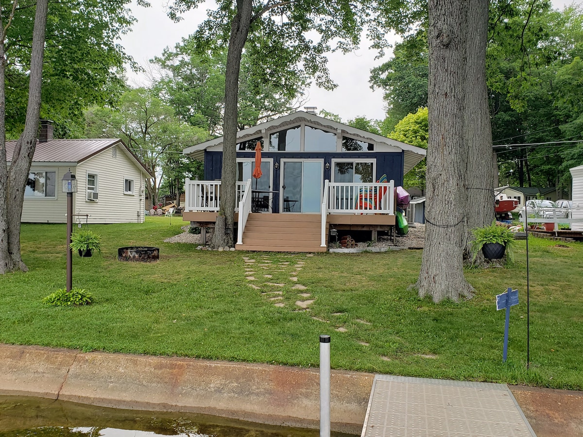 A lakefront cottage is surrounded by greenery and features a large deck with steps leading to a grassy area. The structure has large windows that invite natural light, while a path of stones leads from the dock to the entrance.