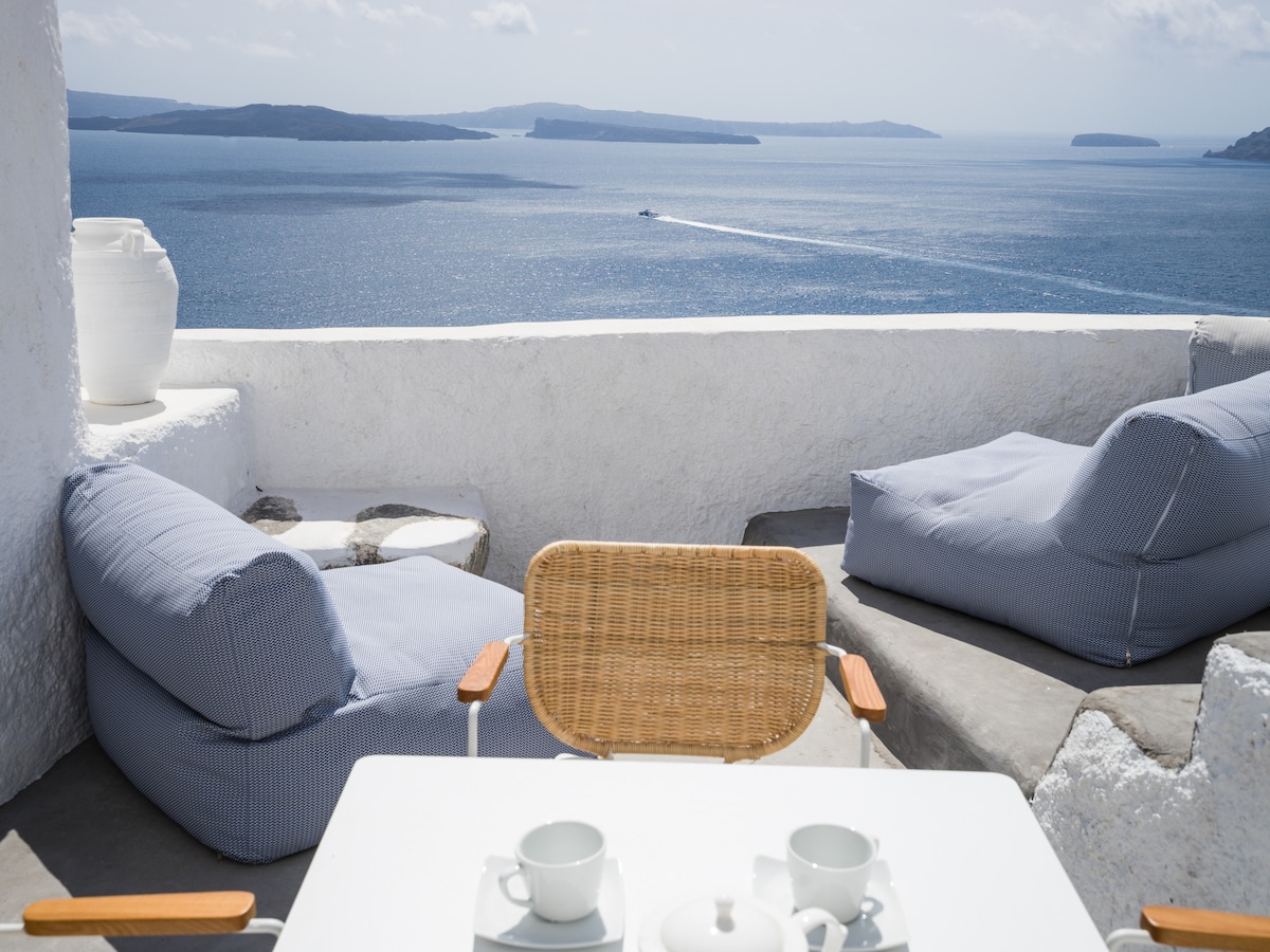 A serene terrace features a small white table set for two, with delicate tea cups and a teapot. Comfortable blue lounge chairs are positioned to overlook stunning sea views, while a large white vase rests nearby. The distant outline of islands can be seen across the water.