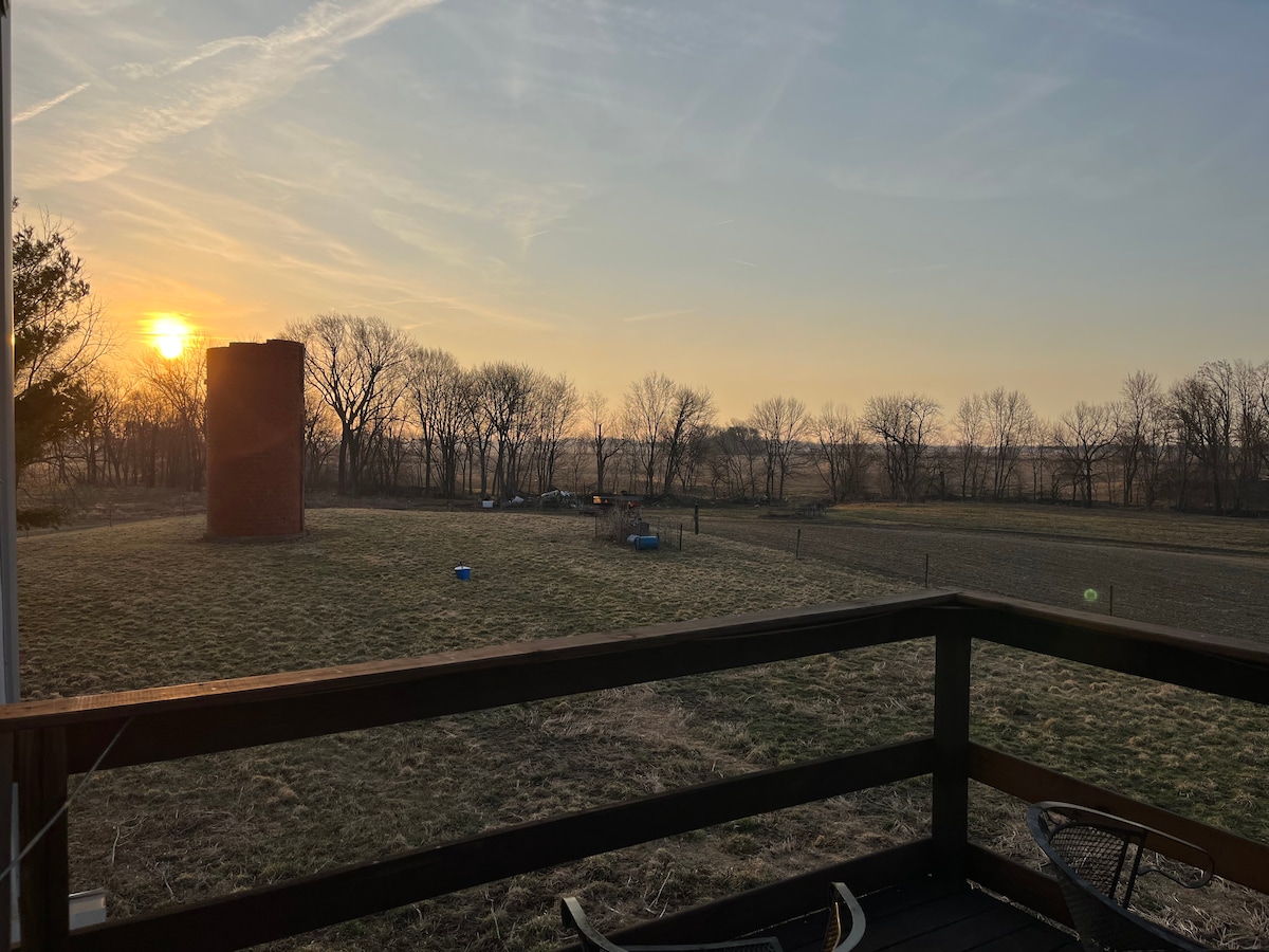 The view from a deck captures a sunset over a tranquil landscape, featuring a red silo in the background and bare trees lining the horizon. The pasture is visible in the foreground, presenting an expansive, peaceful feeling of the countryside.