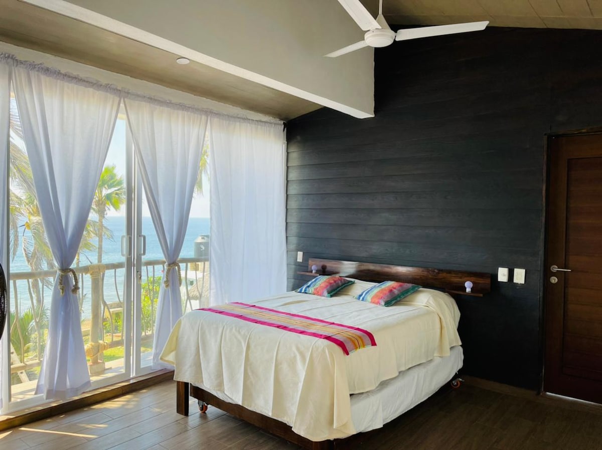 A bright bedroom features large glass doors that open to a scenic view of palm trees and the ocean. A spacious bed is dressed in neutral linens, complemented by colorful decorative pillows. Soft curtains frame the windows, allowing natural light to fill the room.