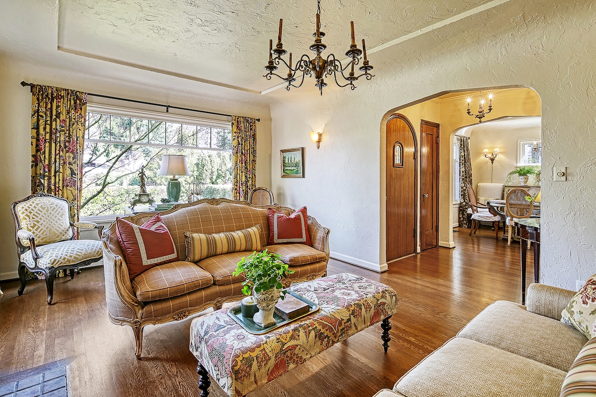 A light-filled living area showcases elegant furnishings, including a patterned ottoman and a vintage sofa. Decorative drapes frame the large window, revealing views of the outside greenery. A dining area is visible through an archway, featuring classic decor and inviting ambiance.