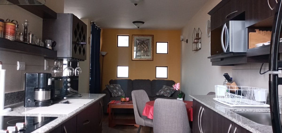 A compact kitchen area features modern appliances, including a coffee maker and stove, with dark cabinetry and granite countertops. A cozy seating area displays a comfortable couch accompanied by a coffee table and dining table set for two. Natural light enters through small square windows.