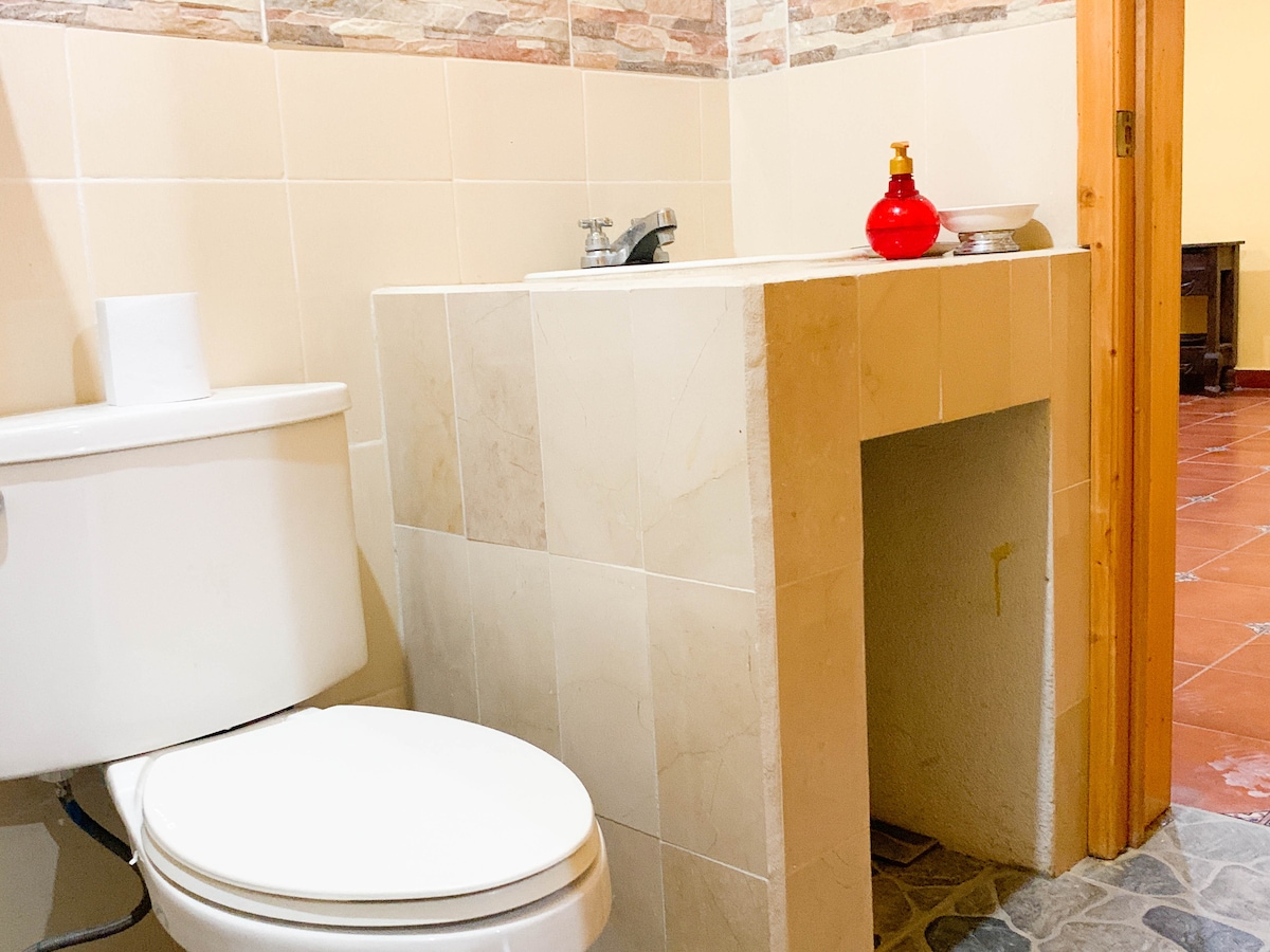 A bathroom is displayed, featuring a white toilet and a sink with a countertop made of light-colored tiles. A red decorative object and a small bowl are positioned on the counter, adding a touch of color to the neutral palette.