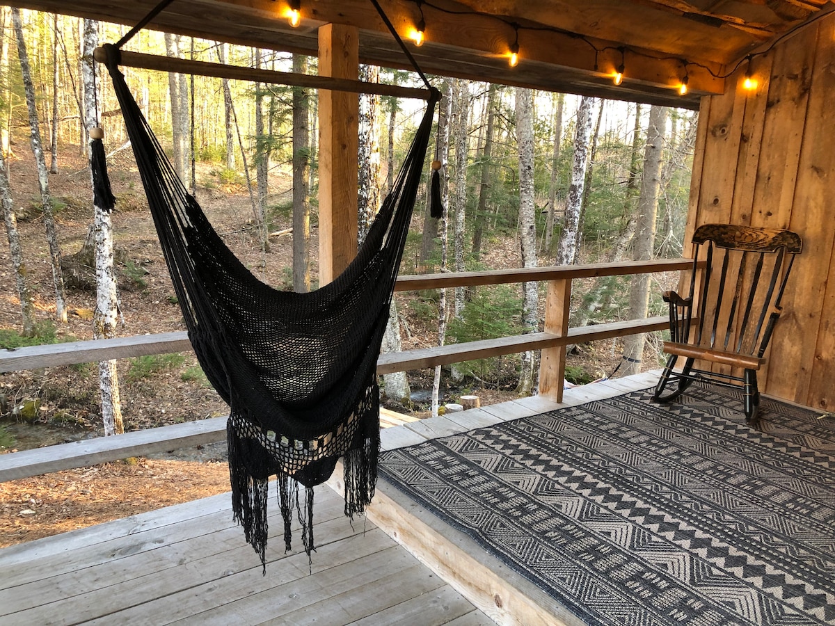 A cozy porch space features a black hammock suspended from the ceiling, inviting relaxation. A wooden rocking chair stands beside it on a patterned area rug, with warm light strings adding a soft glow. A peaceful wooded backdrop frames the scene.
