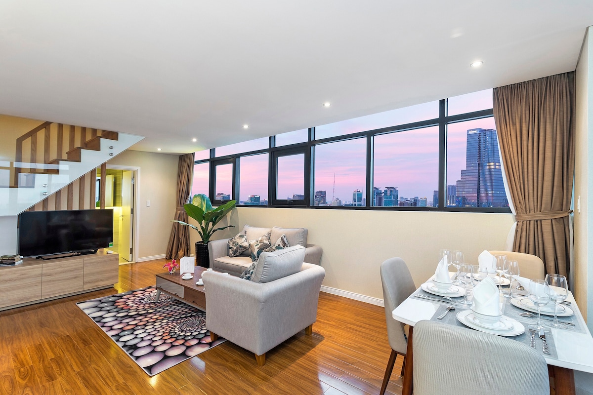 A bright living area features large windows offering city views and an inviting atmosphere. Comfortable seating arrangements, including a sofa and chairs, are complemented by a dining table set for four. The decor includes a vibrant rug and a decorative plant, enhancing the space.