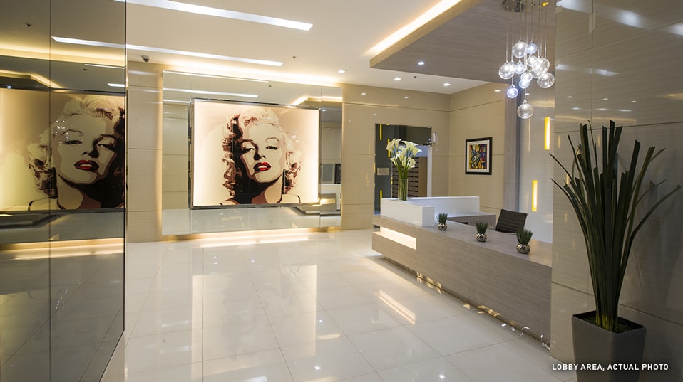 The spacious lobby area features polished white floors and contemporary decor. A reception desk is positioned prominently, complemented by large art pieces on the walls. Elegant lighting fixtures and decorative plants enhance the welcoming ambiance, creating an inviting entrance for guests.