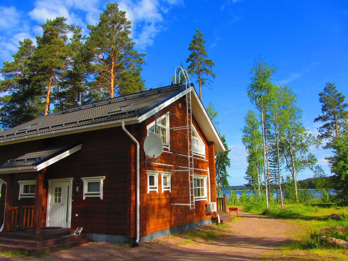 A cottage constructed from natural wood is set against a backdrop of tall pine trees. A clear view of Saimaa Lake is visible in the distance. The design features multiple windows, and a shaded porch area invites relaxation.