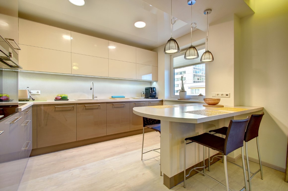 A modern kitchen is equipped with sleek cabinets and a minimalist design. An island with seating is present, accentuated by pendant lighting. Natural light enters through a window, providing a bright atmosphere. Essential appliances and elements like a coffee machine are visible within the space.