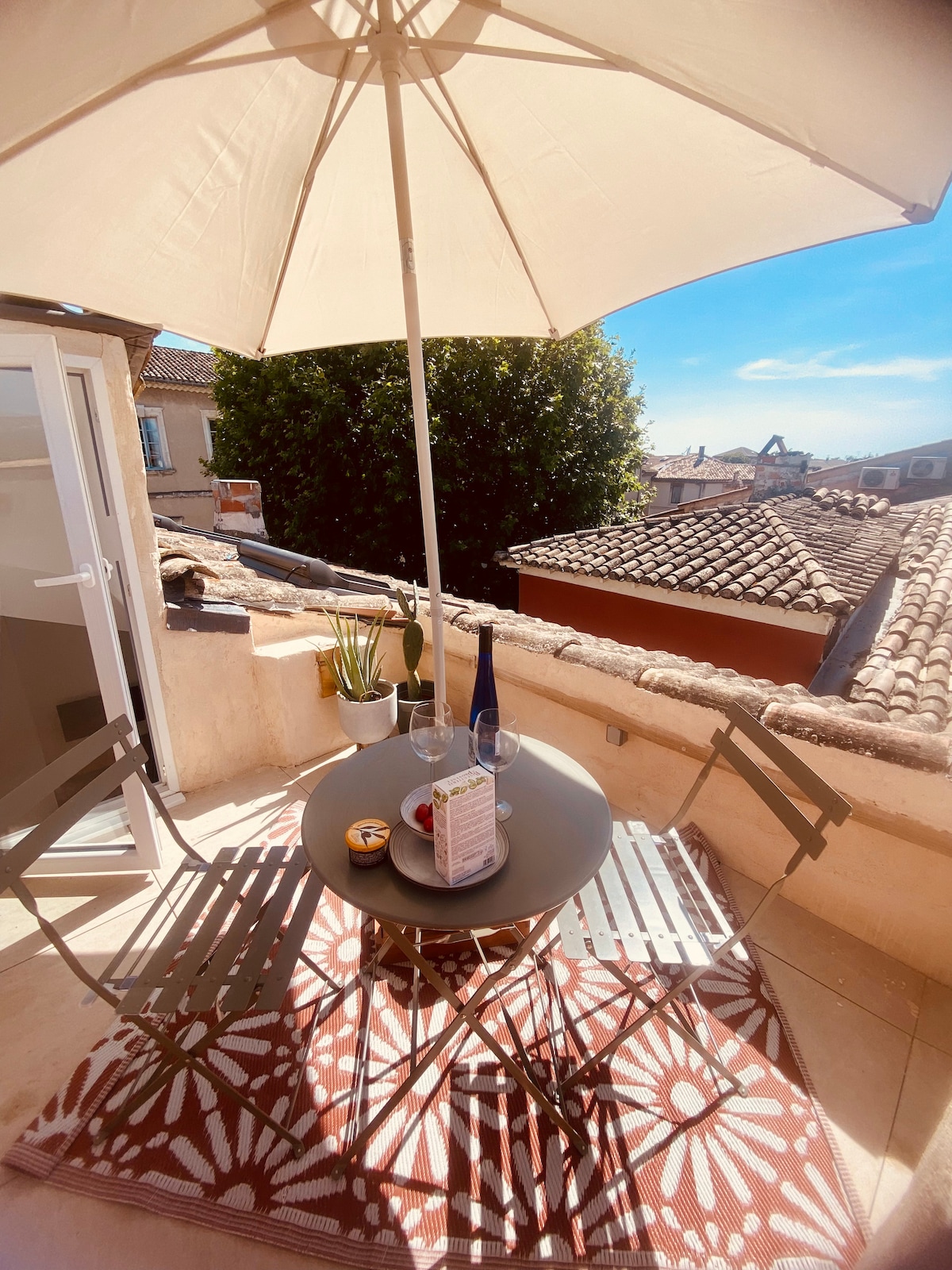 An outdoor terrace features a small round table with two folding chairs beneath a large umbrella, providing shade. A bottle and glasses sit on the table, with a decorative rug and potted plant adding a touch of comfort. Scenic views of the surrounding rooftops are visible.