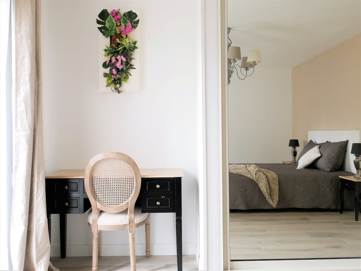 An inviting workspace is provided with a light-colored chair positioned at a black desk, while a vibrant floral wall hanging adds a touch of color. A spacious bed is visible in the background, featuring soft gray bedding and decorative pillows, reflecting a calming atmosphere.