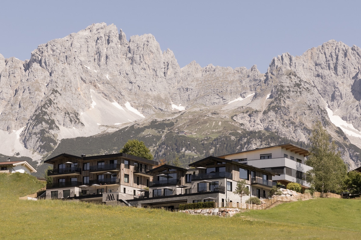 The exterior of the building displays a modern architectural design, set against a backdrop of majestic mountain peaks. The structure features multiple levels with large windows, blending harmoniously with the natural landscape and grassy surroundings.