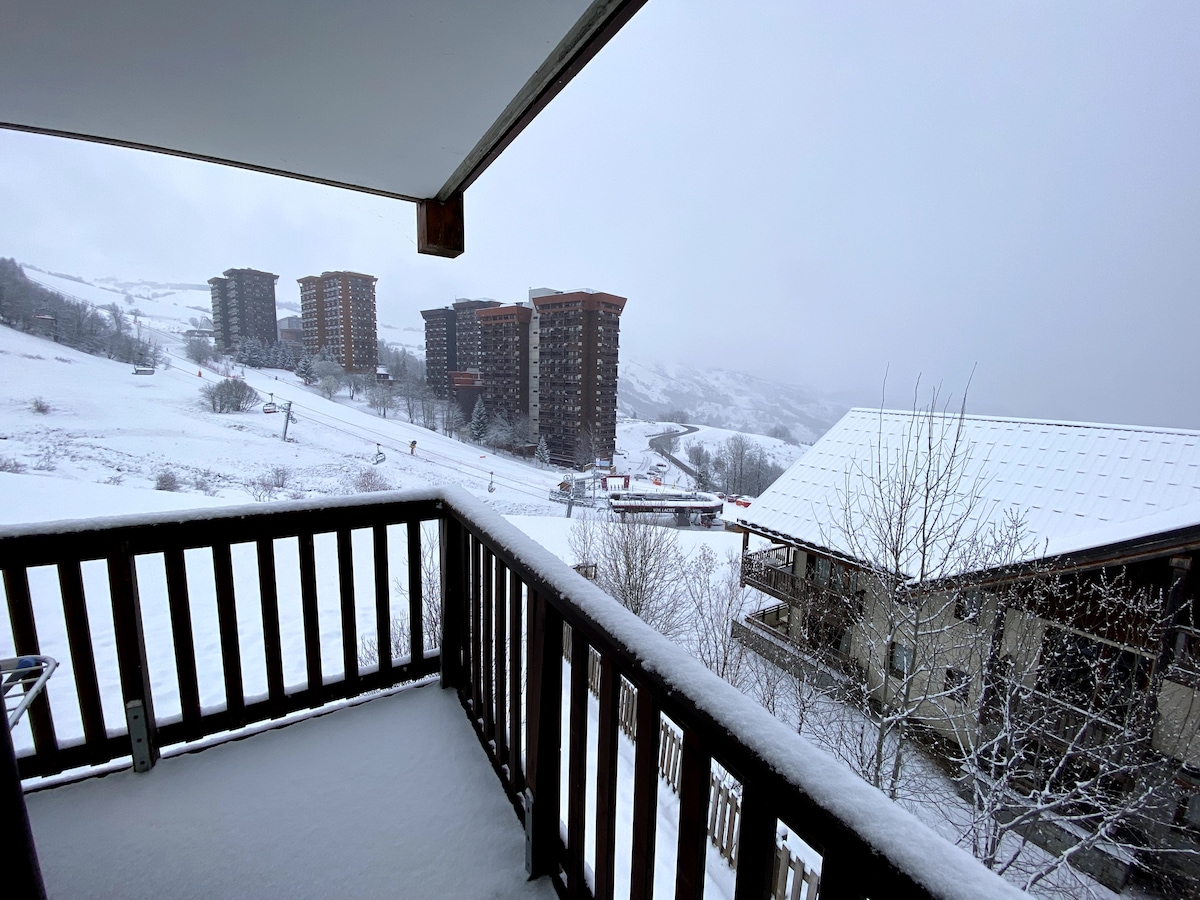 A snow-covered balcony provides a view of the ski area and surrounding buildings. The landscape includes slopes and trees blanketed in white, suggesting a serene winter atmosphere. The design features wooden railings and a clean, spacious floor area.