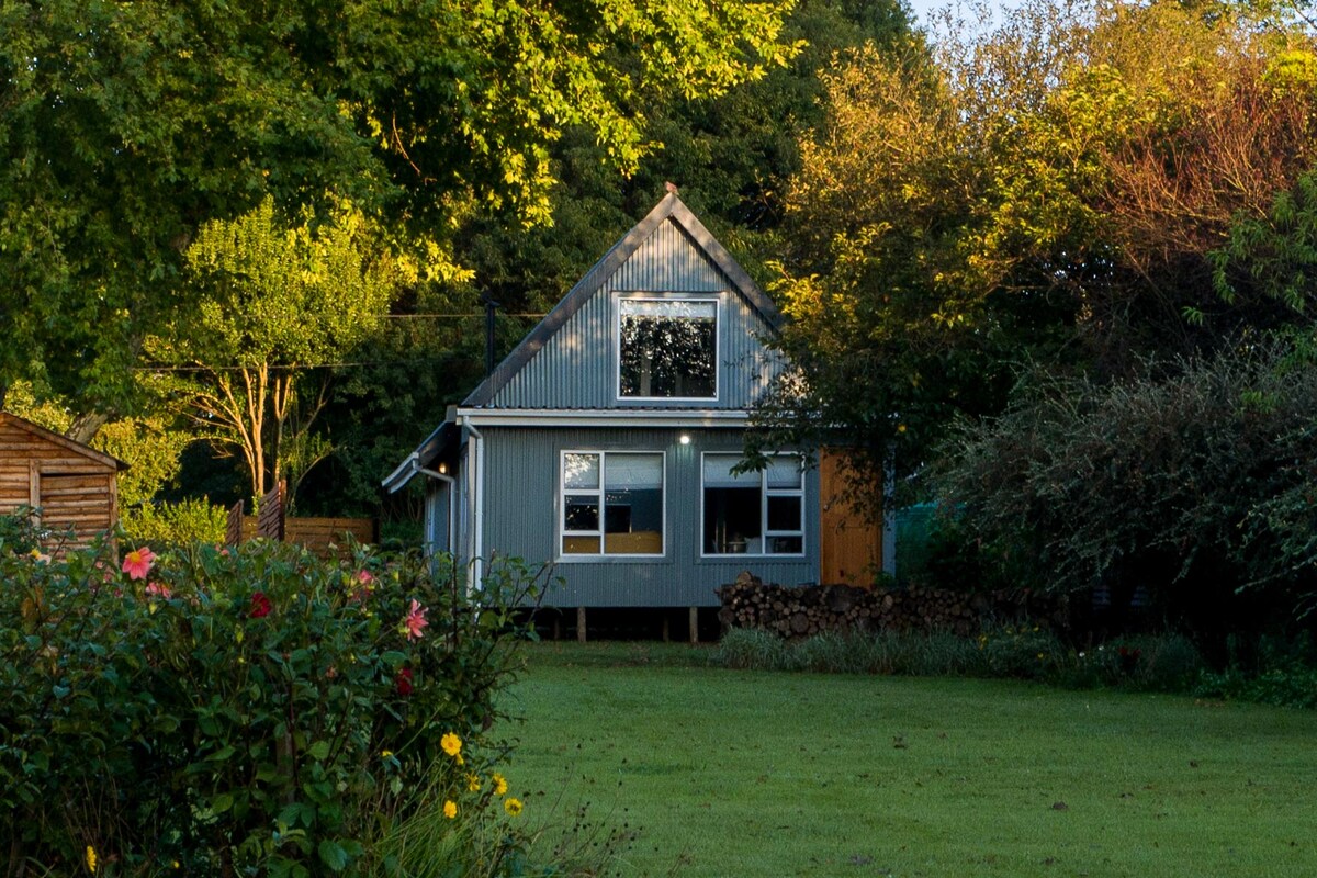 A modern cottage with a grey exterior stands surrounded by lush greenery and colorful flowers. Large windows invite natural light into the space, while a neatly arranged stack of logs adds to the rustic charm. A well-maintained lawn provides a serene setting.