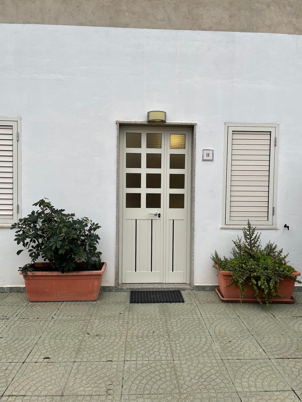 A well-maintained entrance features a pale door with square window panes, flanked by two potted plants. The exterior walls are painted white, and the surrounding tiled area displays a circular pattern. Light filters through the windows, contributing to the inviting entryway.