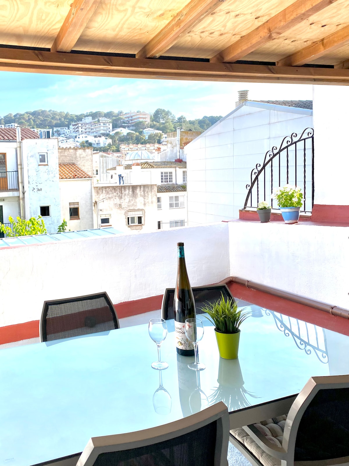 An outdoor terrace features a glass table with four chairs, complemented by a bottle of wine and two glasses. Potted plants add a touch of greenery, while the surrounding buildings and trees are visible in the background, enhancing the tranquil setting.