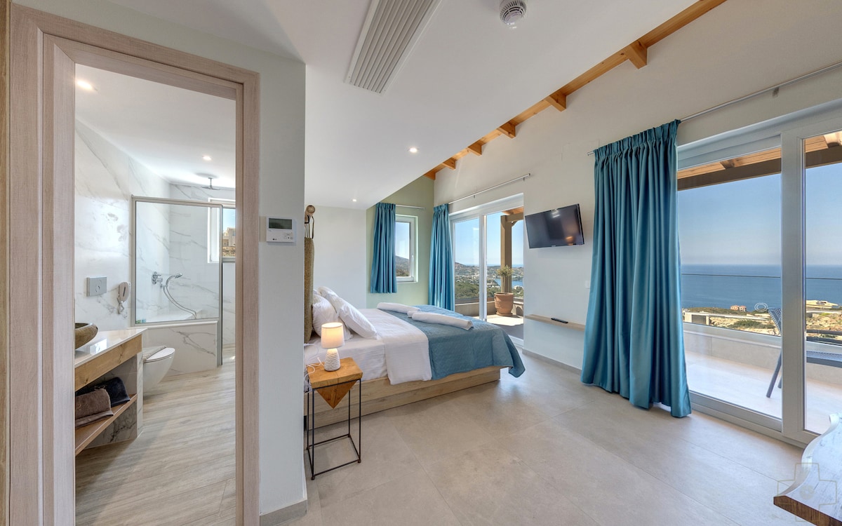 A spacious suite is presented, featuring two beds with soft linens and natural light flooding through large windows. The en-suite bathroom is visible, showcasing modern fixtures. A private balcony offers views of the sea, complemented by soft furnishings in serene earth tones.