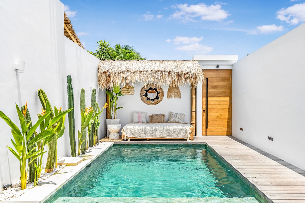 A serene outdoor area features a private pool surrounded by lush green plants. A thatched-roof gazebo offers a cozy seating space with light cushions. The clean white walls reflect natural light, enhancing the inviting ambiance of the space.