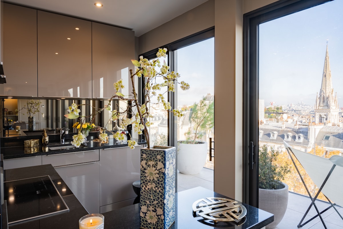 A modern kitchen area features sleek cabinetry and a polished countertop. A decorative vase holds branches with blossoms, enhancing the environment. Large glass windows frame a panoramic view of the city, inviting natural light and showcasing distant architectural spires.