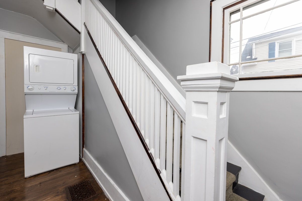 A staircase with wooden handrails is positioned next to a stacked washer and dryer. The walls are painted in neutral grey, and natural light enters through a small window at the top of the steps, illuminating the space.