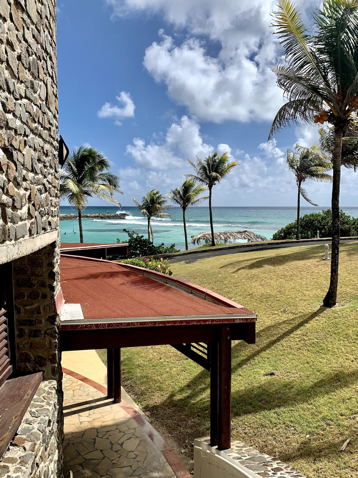 A coastal view reveals the ocean with gentle waves lapping against the shore. Palm trees sway in the breeze, contributing to the tropical ambiance. The stone structure's roofline frames the scene, showcasing the lush greenery of the garden leading down to the beach area.