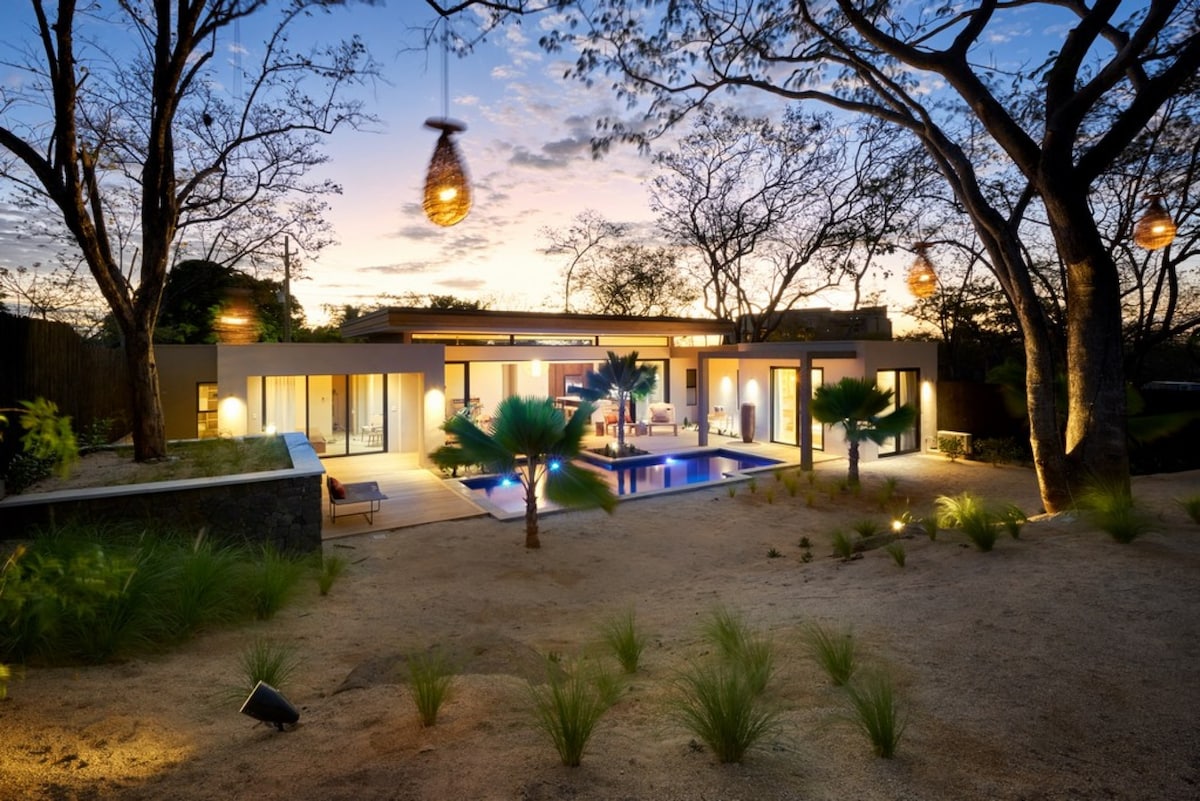 The modern dwelling is framed by trees, with a serene pool reflecting the twilight sky. Soft lighting from lanterns and wall sconces creates a calming ambiance, highlighting the landscaped yard and outdoor seating areas, contributing to a tranquil setting.