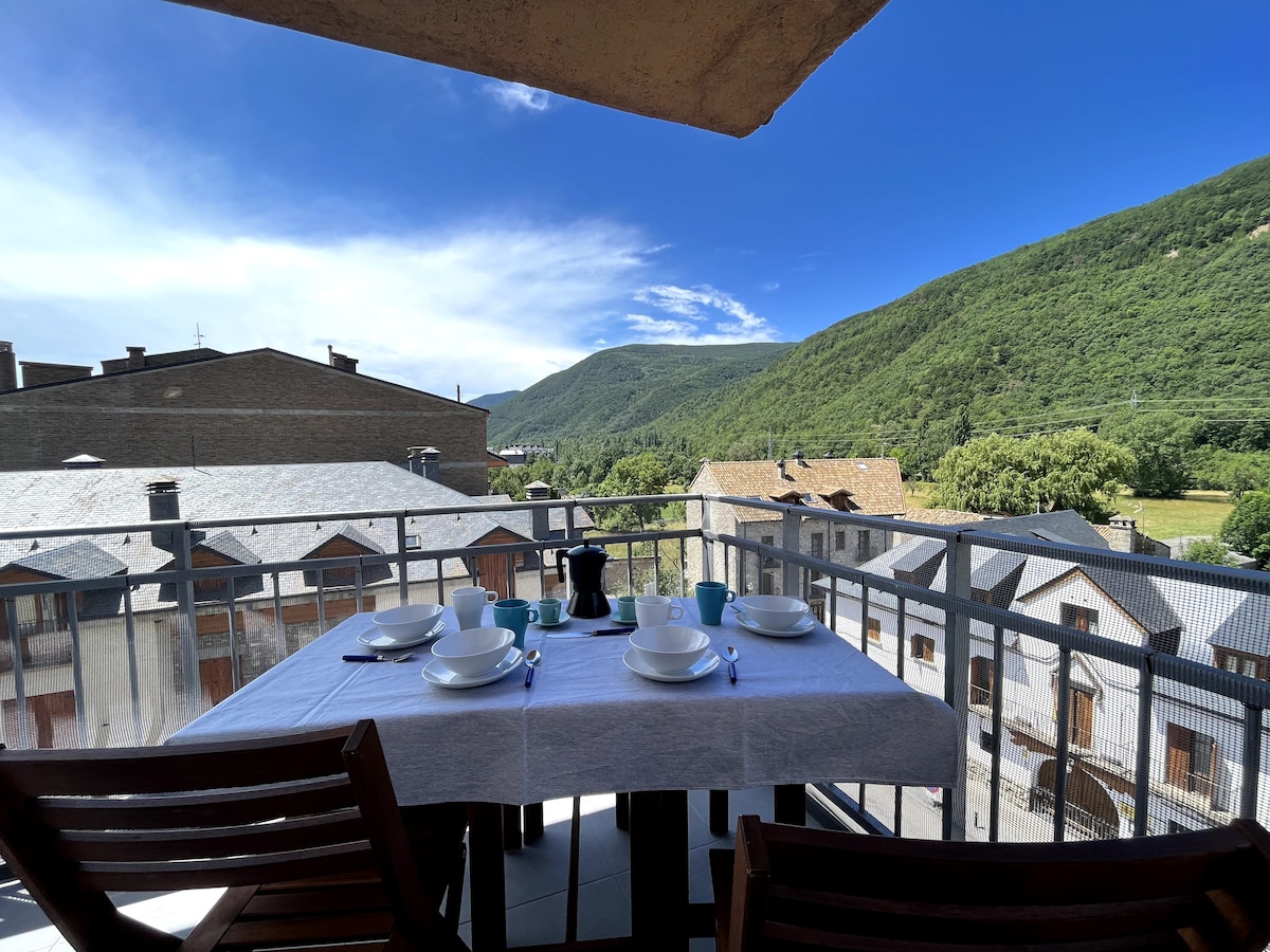 A dining table is set for four on a spacious balcony, overlooking the scenic Tena Valley. Dishes, cups, and a coffee pot are arranged neatly, while the surrounding rooftops and greenery are visible under a bright blue sky.