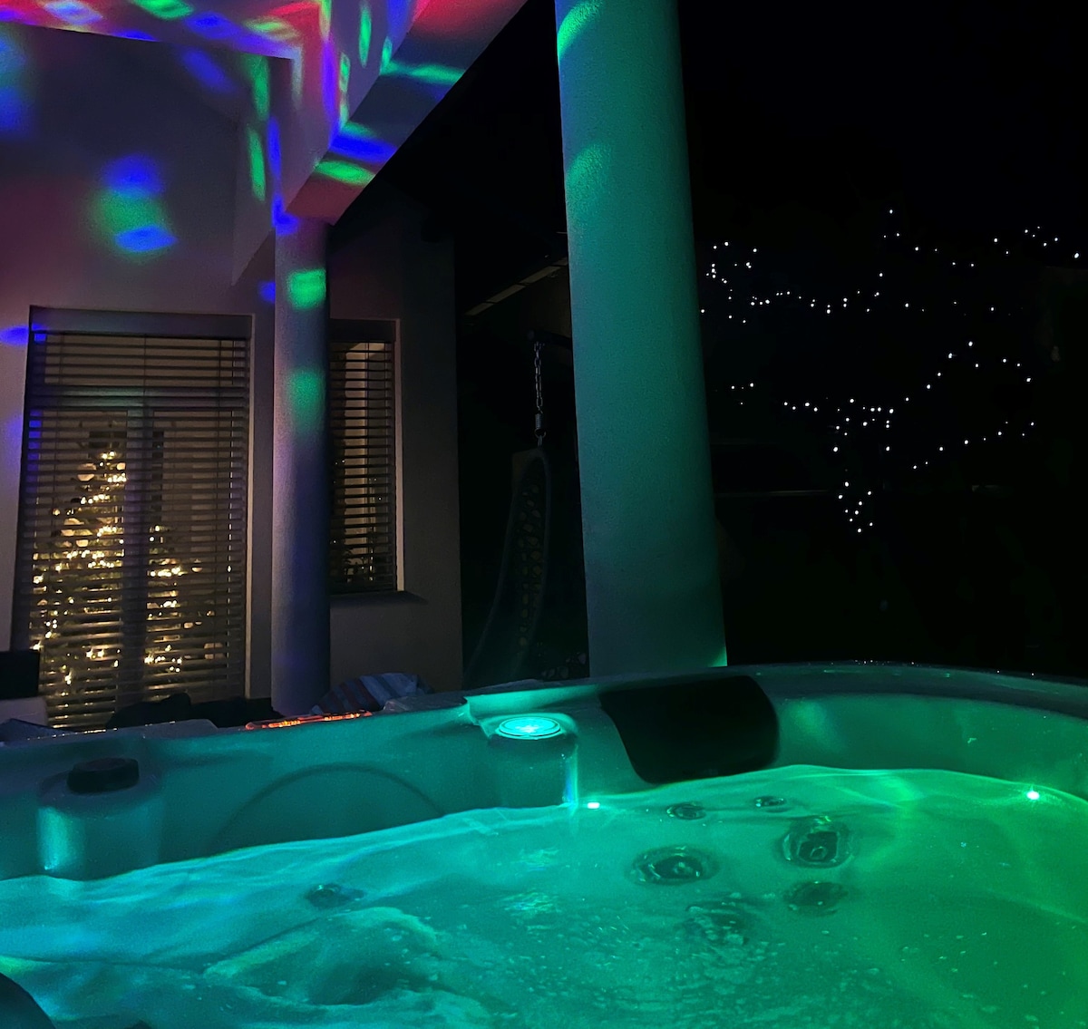 A spacious outdoor Jacuzzi is illuminated with colorful lights, creating a lively ambiance. The structure is surrounded by columns and features a backdrop of soft ambient lighting from nearby greenery, enhancing the atmosphere. Relaxing water jets are visible within the Jacuzzi.