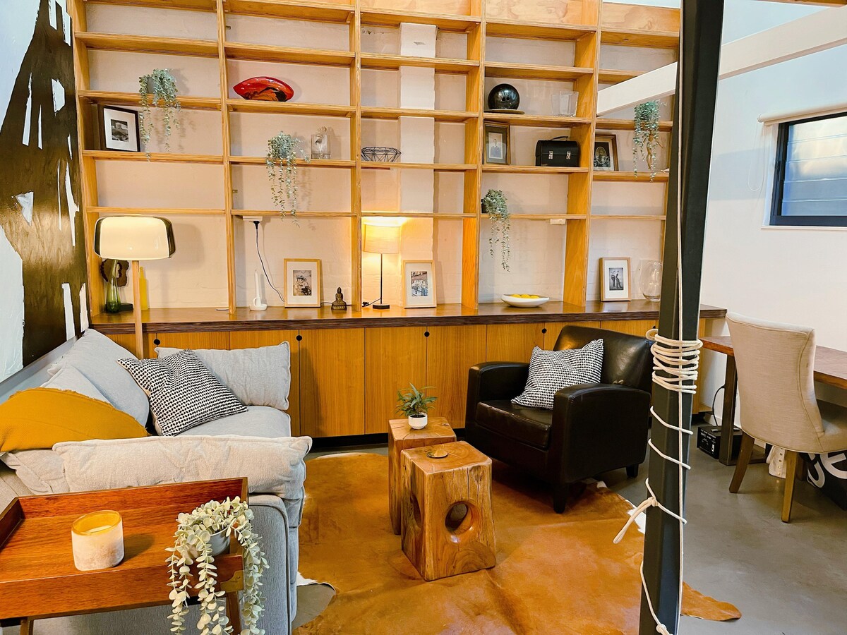A comfortable living area features a cozy sofa and a sleek armchair across from a wooden side table. The wooden shelving unit showcases decorative items and photographs, with subtle lighting enhancing the space. A warm-toned rug is placed on the floor, complementing the modern design.